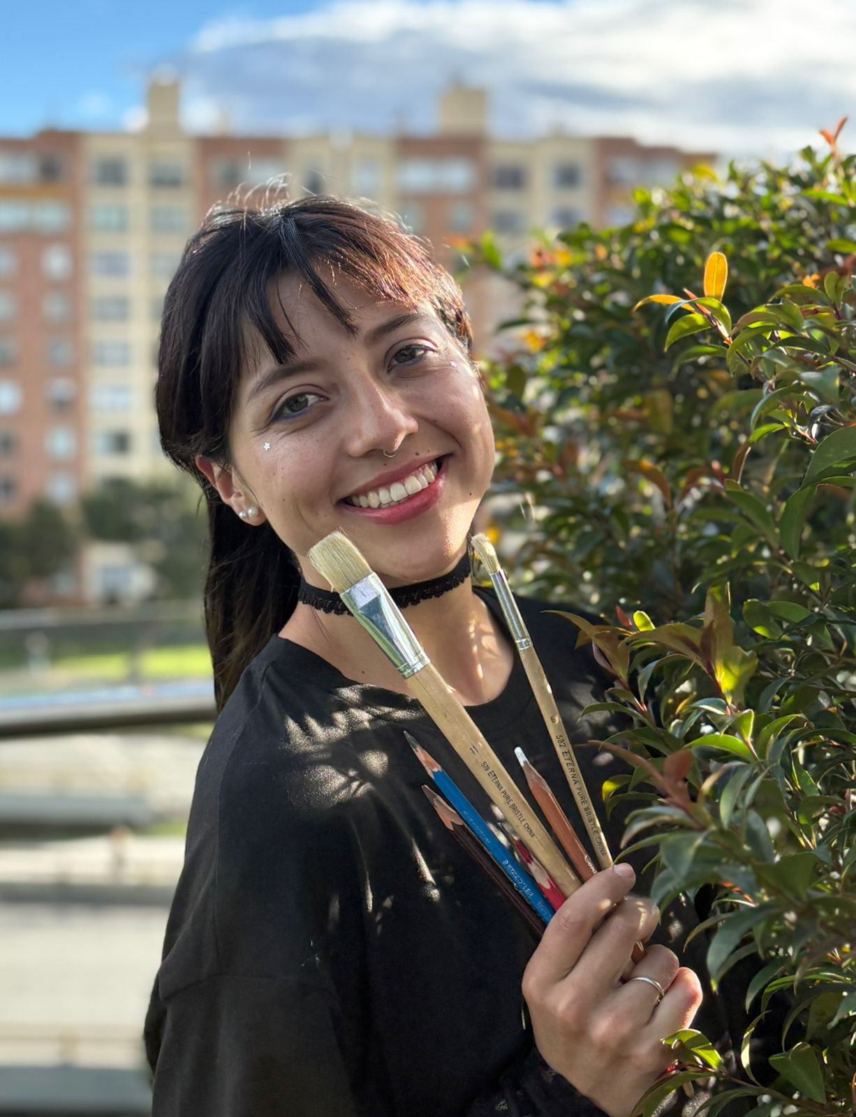 Retrato de bichito. sonriendo al aire libre, con pinceles y lápices de colores en la mano junto a un arbusto.